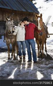 Young adult Caucasian couple kissing and holding horses with stable in background.