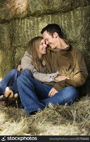 Young adult Caucasian couple sitting on hay hugging and smiling at each other.