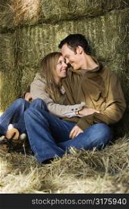 Young adult Caucasian couple sitting on hay hugging and smiling at each other.