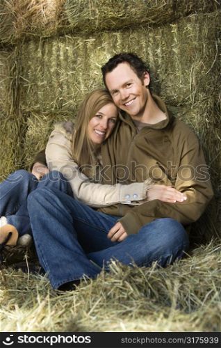 Young adult Caucasian couple sitting on hay hugging and smiling at viewer.