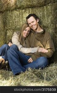 Young adult Caucasian couple sitting on hay hugging and smiling at viewer.