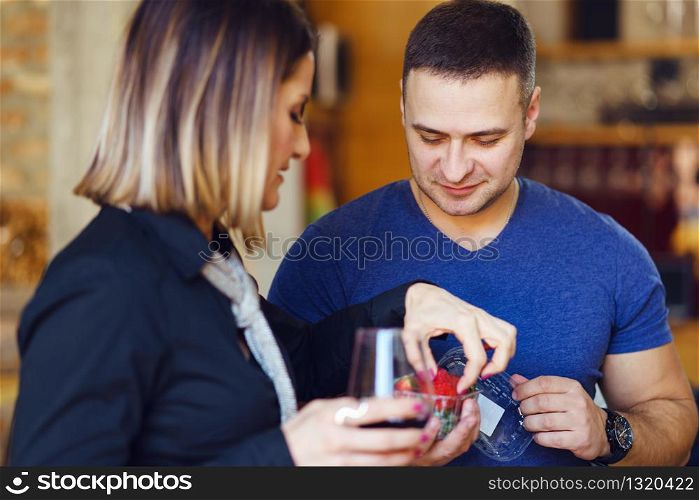Young adult caucasian man and woman holding glass of wine offering strawberries wearing blue shirt happy side view at home party day