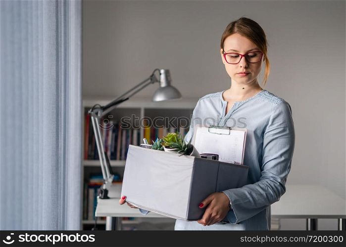 Young adult caucasian woman female girl standing by the window at the office losing her job holding personal items things in box being fired from work dismissed due to crisis recession quitting