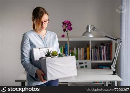Young adult caucasian woman female girl standing in front of table at the office losing her job holding personal items things in box being fired from work dismissed due to crisis recession quitting