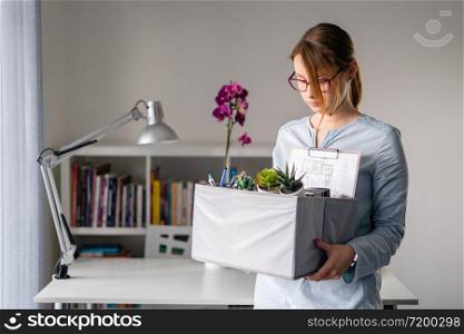 Young adult caucasian woman female girl standing in front of table at the office losing her job holding personal items things in box being fired from work dismissed due to crisis recession quitting