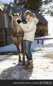 Young adult Caucasian woman petting horse with stable in background in winter.