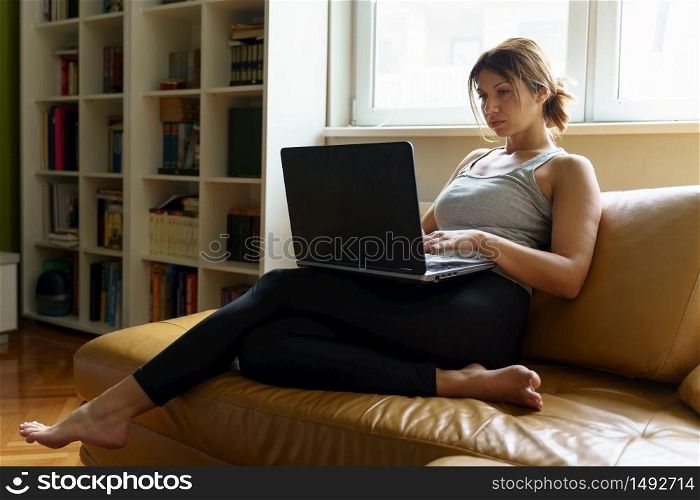 Young adult caucasian woman working on the laptop computer from home sitting on the sofa bed in day taping online female freelancer or shopping online efficiency and expertise making money concept