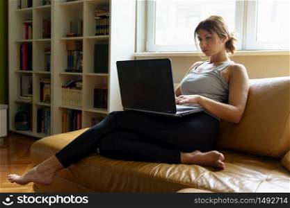 Young adult caucasian woman working on the laptop computer from home sitting on the sofa bed in day taping online female freelancer or shopping online efficiency and expertise making money concept