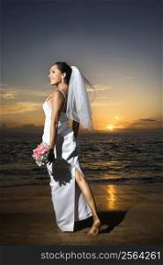Young adult female Caucasian bride standing holding bouquet on beach.