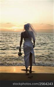 Young adult female Caucasian bride standing on beach at sunset.