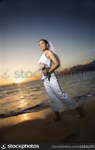 Young adult female Caucasian bride walking holding bouquet on beach.