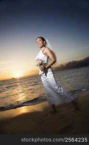 Young adult female Caucasian bride walking holding bouquet on beach.