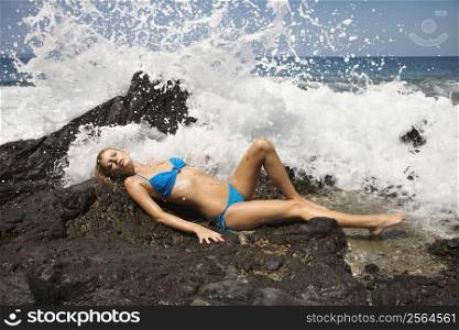 Young adult female Caucasian lying on rock in bikini with wave crashing.