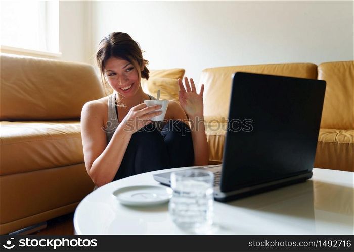 Young adult female caucasian woman sitting by the sofa bed at home using laptop computer to connect and contact her friends online using communication platform or social network waving and smiling