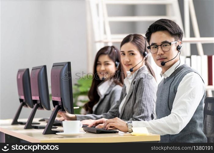 Young adult friendly and confidence operator asian man agent with headsets working in a call center with his colleague team working as customer service and technical support workplace in background.