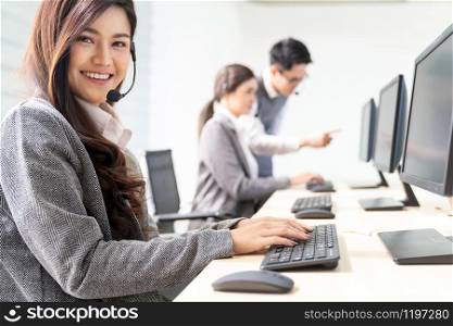 Young adult friendly and confidence operator woman agent smiling with headsets working in a call center with her colleague team working as customer service and technical support workplace in background.