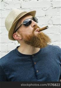 Young adult male wearing fedora and sunglasses smoking a cigar on the street.