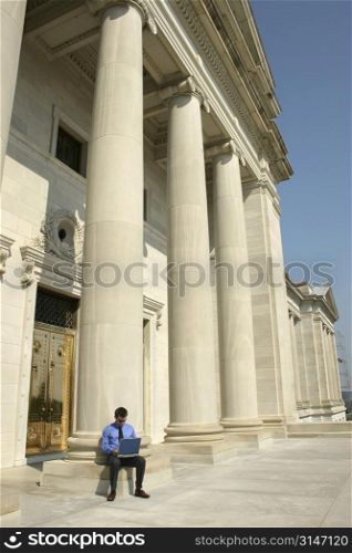 Young adult male working on a laptop computer in front of a government building.