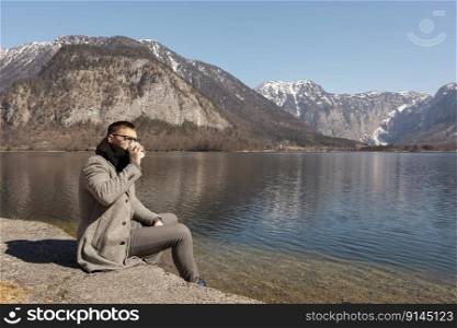 Young adult man sitting outdoors, drinking coffee and enjoying mountains, lake, good weather, blue sky and sun. Beautiful landscape. Time with yourself, dreaming, relaxation, mental health. Holiday. Young adult man sitting outdoors, drinking coffee and enjoying mountains, lake, good weather, blue sky and sun. Beautiful landscape. Time with yourself, dreaming, relaxation, mental health. Holiday.