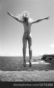 Young adult nude Caucasian woman jumping in air near cliff at Maui coast.