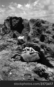 Young adult nude Caucasian woman lying on boulder in rocky Maui landscape.