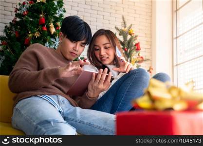 Young Adult teenager couple using tablet on sofa in living room for online shopping with gift box with Christmas tree in background