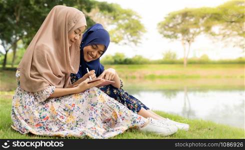 Young adult teenager Muslim Islamic Thai Asian women using smartphones and digital tablet in social media beside lake and green park in University college school.