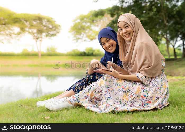 Young adult teenager Muslim Islamic Thai Asian women using smartphones and digital tablet in social media beside lake and green park in University college school.