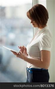 Young adult woman with short hair using tablet while standing at window.. Side view of adult woman using tablet