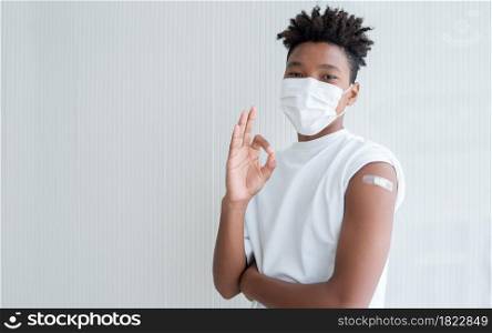 Young African American boy wear face mask smile and showing plaster stuck on arm to show that man get vaccinated with hand make okay sign. White background. Copy space