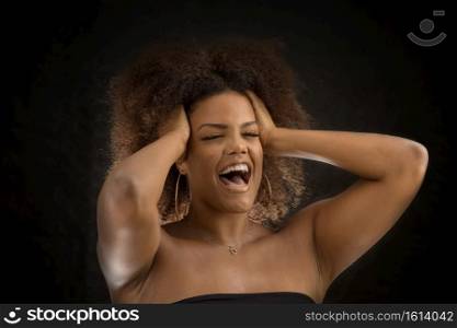 Young African American female with hands on head yelling in madness with eyes closed on black background. Black woman squeezing head and screaming in crisis