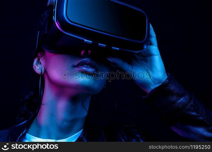 Young african american girl playing game using VR glasses, enjoying 360 degree virtual reality headset for gaming, isolated on black background in neon light