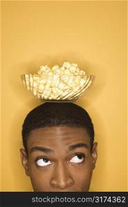 Young African-American man balancing bowl of popcorn on his head on yellow background.