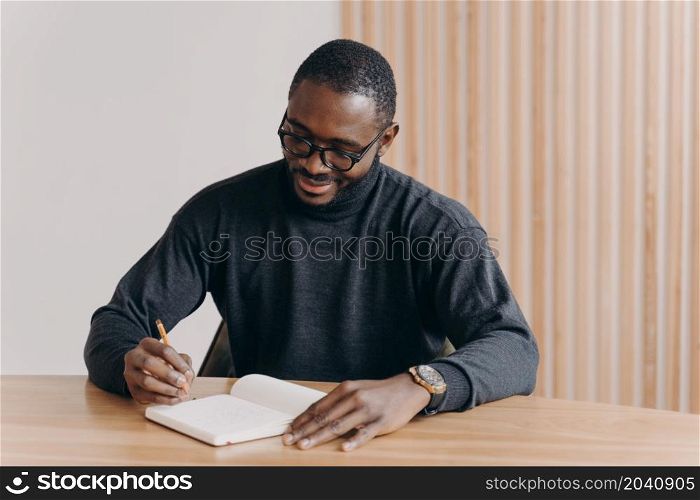 Young african american man entrepreneur in glasses taking notes in agenda while sitting at desk at home or office,planning workday, writing down some fresh ideas or work schedule. Young african american man entrepreneur in glasses taking notes in agenda