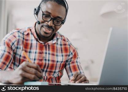 Young african american man in headset has video chat on laptop. Student gets distant education and taking notes. Remote lesson at college. Friendly manager working from home. Client support service.. African american man in headset has video chat on laptop. Remote lesson. Client support service.