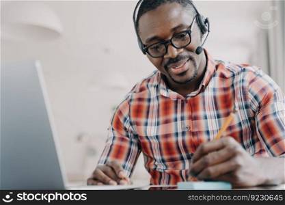 Young african american man in headset has video chat on laptop. Student gets distant education and taking notes. Remote lesson at college. Friendly manager working from home. Client support service.. African american man in headset has video chat on laptop. Remote lesson. Client support service.