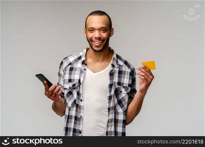 Young african-american man smiling and hoding credit card and mobile cell phone over grey background with copy space.. Young african-american man smiling and hoding credit card and mobile cell phone over grey background with copy space