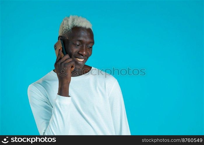 Young african american man speaks with smile on phone. Guy holding and using smart phone. Blue studio background. Young african american man speaks with smile on phone. Guy holding and using smart phone. Blue studio background.
