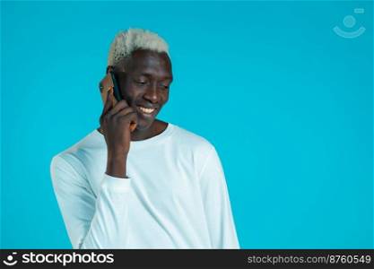 Young african american man speaks with smile on phone. Guy holding and using smart phone. Blue studio background. Young african american man speaks with smile on phone. Guy holding and using smart phone. Blue studio background.