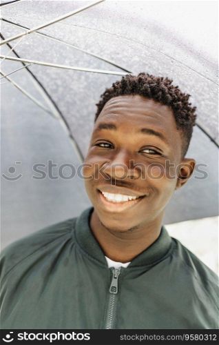 Young African American man with umbrella, under rain, smiling on street outdoor. Fall or spring season. Young African American man under black umbrella in rain, smiling. Fall or spring weather
