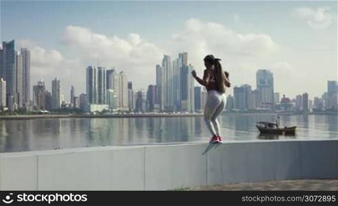Young african american woman in sports clothing, exercising early morning in the city and jumping for joy at the end of her daily workout. Buildings and urban skyline in background. Slowmotion 120p