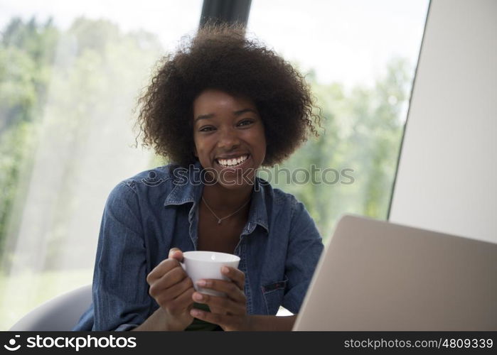 Young african american woman smiling sitting near bright window while looking at open laptop computer on table and holding white mug in her luxury home