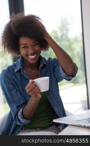 Young african american woman smiling sitting near bright window while looking at open laptop computer on table and holding white mug in her luxury home