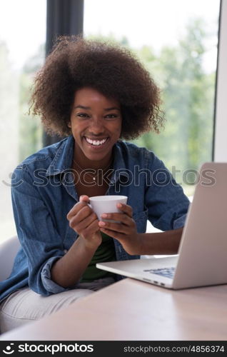 Young african american woman smiling sitting near bright window while looking at open laptop computer on table and holding white mug in her luxury home