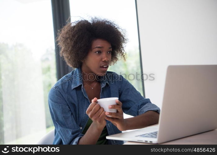 Young african american woman smiling sitting near bright window while looking at open laptop computer on table and holding white mug in her luxury home