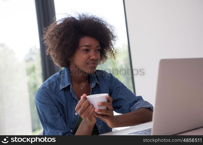 Young african american woman smiling sitting near bright window while looking at open laptop computer on table and holding white mug in her luxury home