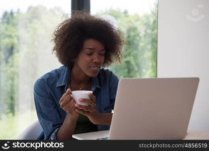 Young african american woman smiling sitting near bright window while looking at open laptop computer on table and holding white mug in her luxury home