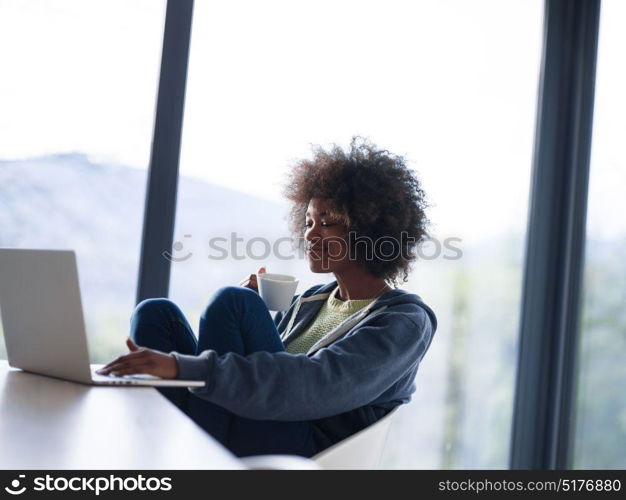 Young african american woman smiling sitting near bright window while looking at open laptop computer on table and holding white mug in her luxury home