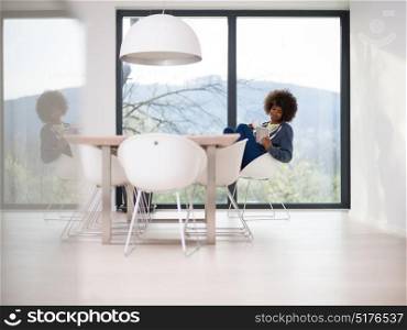 Young african american woman smiling sitting near bright window while looking tablet computer and holding white mug in her luxury home
