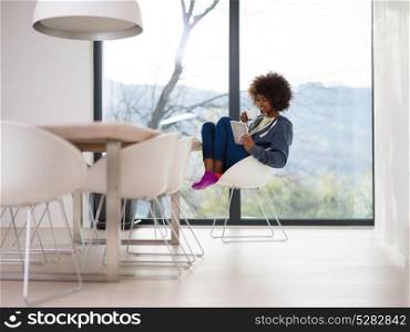 Young african american woman smiling sitting near bright window while looking tablet computer and holding white mug in her luxury home